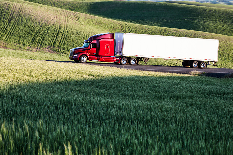 Semi truck with a red cab driving down a road with fields on both sides